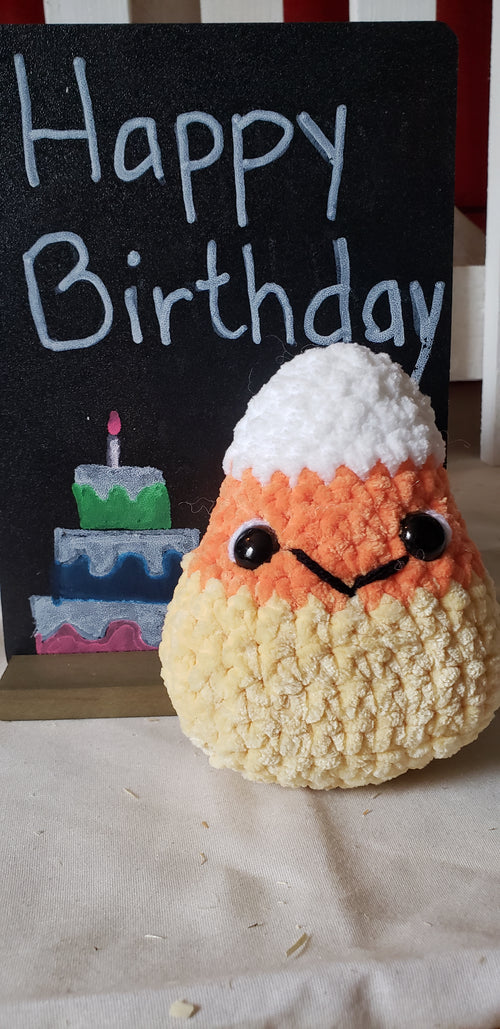 Crochet candy corn plushie with smiley face next to Happy Birthday chalkboard sign - front view
