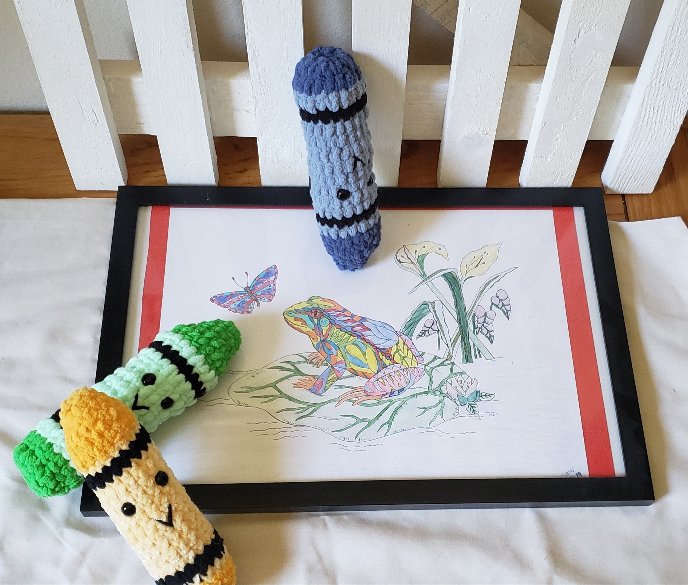 Handmade crochet plush crayons in blue and green/yellow/white with sweet faces, displayed next to a colorful coloring page