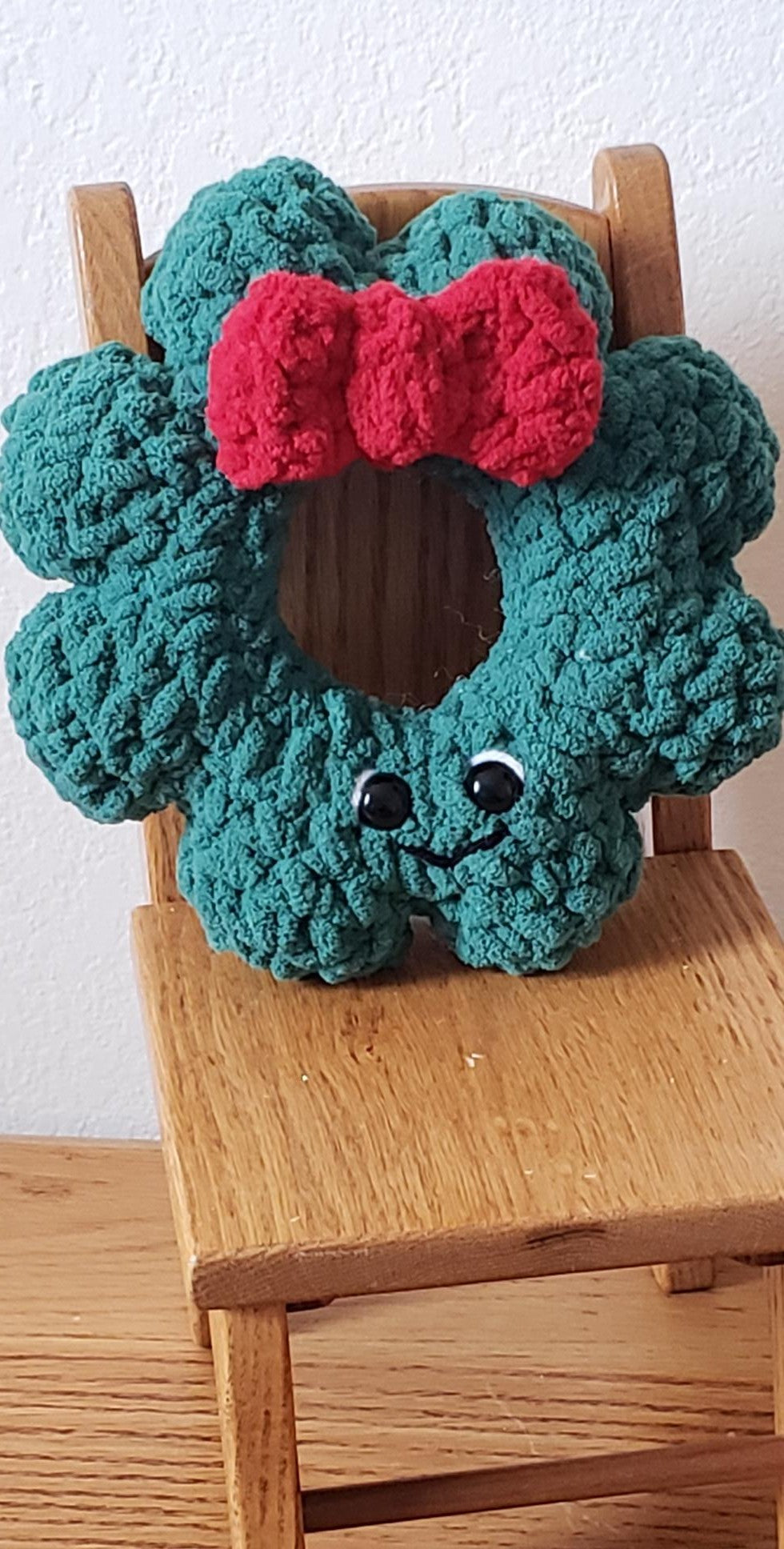 Crochet plush mini Christmas wreath with red bow and smiley face displayed on small wooden chair - front view