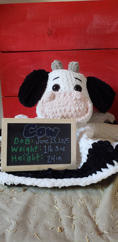 Handmade crochet cow lovey blanket with black and white stuffed head and arms, holding a chalkboard birth announcement sign