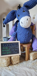 Blue and lavender crocheted donkey plushie sitting on hay bales with a chalkboard birth announcement sign
