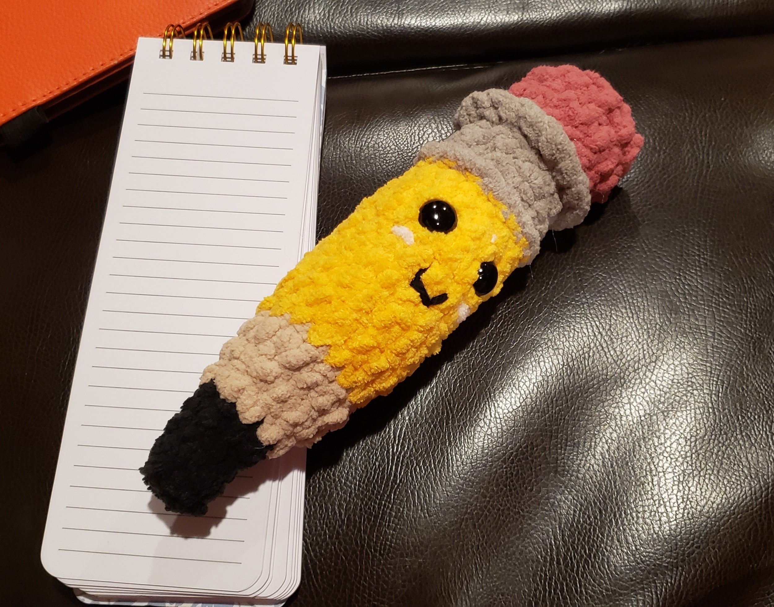Handmade crochet pencil plushie with happy face, yellow body, gray ferrule, and pink eraser, approximately 9 inches long