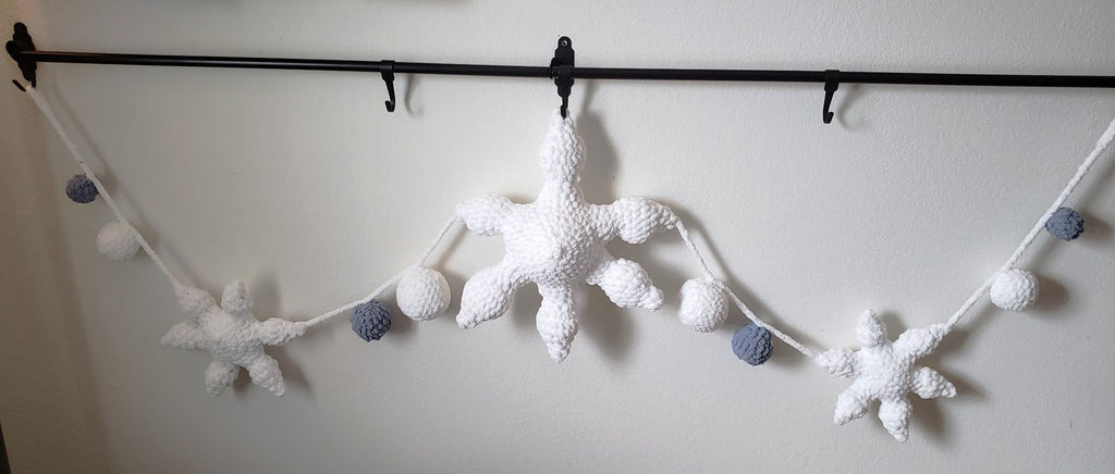Crochet plush snowflake garland full length view showing 1 large center snowflake, 2 mini snowflakes and 8 plush balls in white and slate blue